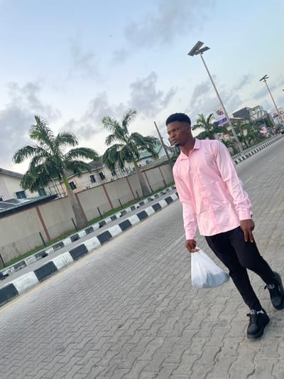 A young man in a pink shirt, black pants, and black shoes stands on a paved walkway, holding a white plastic bag. Palm trees line the street and a multi-story building can be seen in the background. The sky is overcast. The location is Ilado, Nigeria, as specified.