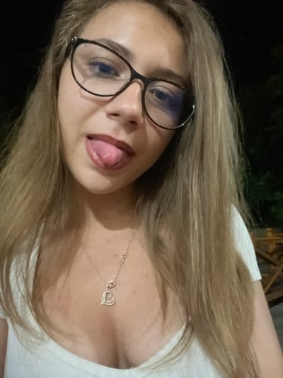 A young woman with blonde hair and glasses is sticking out her tongue. She is wearing a white top and a silver necklace with a heart-shaped pendant. The background is dark, with some trees visible. The scene appears to be outdoors at night. There is no indication of the location being in Beguntsi, Bulgaria.