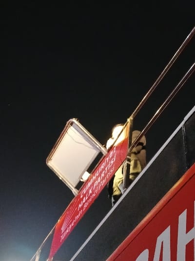 A person is standing on a double-decker bus in Istanbul, Türkiye (latitude: 41.011, longitude: 28.964). The bus has a large red sign on the side that reads "CUMHURİYETİ HALK PARTİSİ" and "Türkiye'nin Birinci Partisi". There is a white light above the person. The bus is facing forward, and it is unclear where the person is looking. The environment is dark, with only the lights from the bus and the city in the distance illuminating the scene. The person is wearing a light-colored shirt and dark pants. They are obscured by the sign and the bus. The bus is brightly lit, and the scene is taken from a low angle, looking up at the bus. It is nighttime.