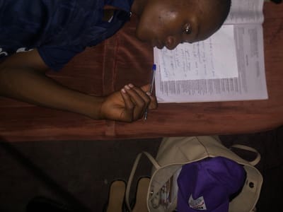 A young Black man is lying on a brown couch, possibly in a home in Atani, Nigeria. He is wearing a dark blue shirt. He is holding a blue pen in his right hand and appears to be studying a textbook and notes on a white sheet of paper. The text in the book and on the notes is partially legible. A beige purse with a purple hat inside is visible near the top left of the image. Sandals are near the purse. The lighting is dim.