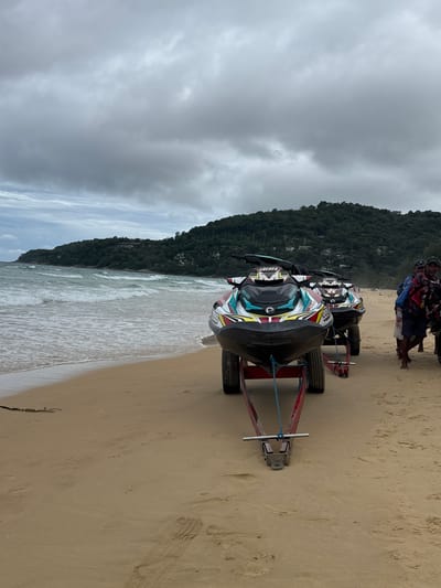Two jet skis on red trailers are parked on a sandy beach in Karon, Thailand. The jet ski in the foreground has a predominantly white, red, and teal color scheme with the number "399" visible on its front. The jet ski behind it shares a similar design. The beach is lined with small waves breaking on the shore, and a lush, green, tree-covered hillside with some buildings is visible in the background. The sky is overcast and cloudy. Three men are standing on the beach to the right of the jet skis; one is looking at the camera and smiling. The sand is imprinted with footprints and tire tracks. The scene suggests a recreational area preparing for or recovering from water activities.