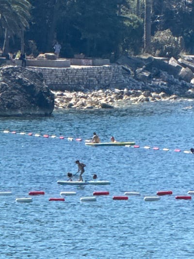 The image depicts a sunny day at Kamenovo, Montenegro, with a focus on a rocky shoreline transitioning into blue water. In the water, several individuals are engaged in activities related to kayaking and paddleboarding. One person is seated in a kayak, while another is on a paddleboard, both appearing to be recreational participants. A buoy line with alternating red and white markers stretches across the water, delineating a swimming or activity area. The background shows lush green trees and vegetation on the hillside, suggesting a warm climate. The lighting indicates it's likely midday, with bright sunlight casting shadows and highlighting the textures of the rocks and water. The overall scene conveys a sense of leisure and outdoor recreation.