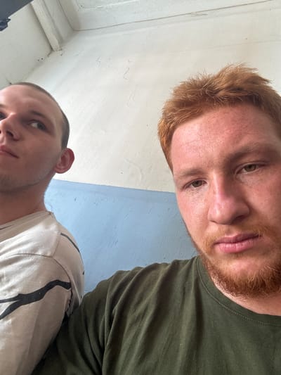 The image is a close-up selfie featuring two men in what appears to be an indoor setting with a light-colored wall and a blue-painted section. The man in the foreground has reddish-brown hair and beard, with fair skin and green eyes. He is wearing a dark green t-shirt. The man in the background has light brown hair and is looking to his left. He is wearing a light-colored t-shirt with a black pattern. The background wall shows some signs of wear and tear. There are no explicit details in the image to suggest a specific location within Makiivka, Ukraine.