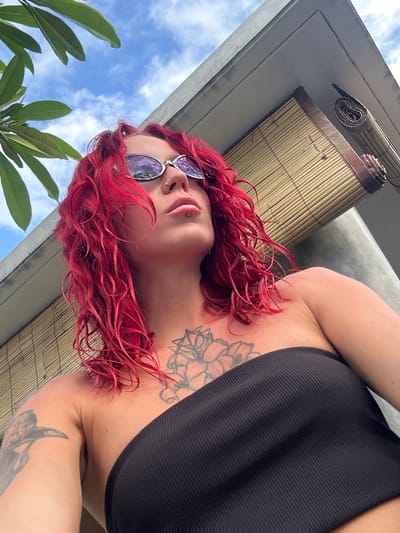 A medium, upward-facing shot in Dalung, Indonesia shows a fair-skinned person with red, curly shoulder-length hair and purple-tinted sunglasses. The person has a lip piercing, and tattoos are visible on the chest and arm. The person is wearing a black, ribbed top. A partially rolled up bamboo sunshade is positioned above them. A tree with green leaves is located in the lower-left corner of the frame, and a blue sky with white clouds is visible above the tree and behind the person.