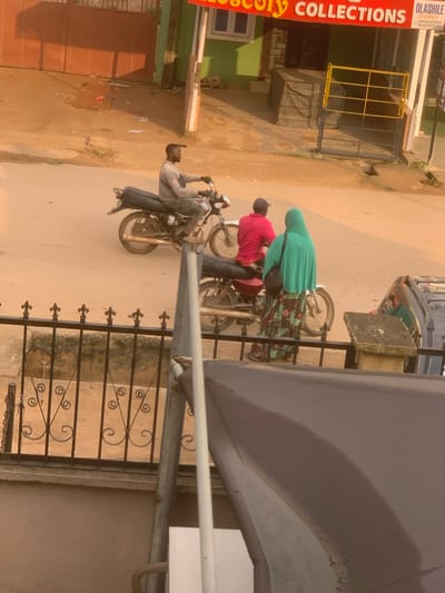 A street scene in Shagamu, Nigeria, features a man riding a motorcycle with a passenger behind him. The man wears a gray long-sleeved shirt and dark pants. The passenger, a woman, is dressed in a vibrant green hijab and a patterned dress. They are on a paved road. To the left of the motorcycles, a brown building with a red-brown gate is visible, possibly a shop or residential building. Further back, a green building with a sign reading "OLASHILE PAINT" and a phone number can be seen. A yellow metal structure, possibly a fence or barricade, stands near the green building. In the foreground, a black wrought-iron fence runs horizontally across the bottom of the image. The ground appears to be a mixture of dirt and asphalt.