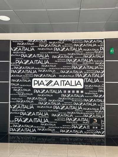 The image displays a vertical section of a wall or partition in an indoor setting, adorned with a repeating textual pattern. The primary text, "PIAZZA ITALIA," is rendered in white against a black background, appearing in various sizes, font styles, and orientations, creating a textured, almost wallpaper-like effect.
In a central area, a more prominent white-boxed "ITALIA" is visible, alongside "PIAZZA." This section also features a logo for "ALTAHAN" with Arabic text, "شركة عدنان الطحان وأولاده التجارية" (Adnan Al-Tahan and Sons Trading Company), and the website "www.altahan.com." Below this branding, social media icons for Facebook, YouTube, Pinterest, Instagram, and Twitter are present.
A small, bright green rectangular tag bearing the number "11" is affixed to the wall in the upper left. The wall surface to the left of the patterned area is a plain grey, appearing as segmented panels. To the right, reflections of the "PIAZZA ITALIA" pattern are visible on a mirrored surface, showing the text in reverse. No people or specific activities are depicted. The scene suggests a commercial or public interior within Kafr Sousa Municipality, Syria.