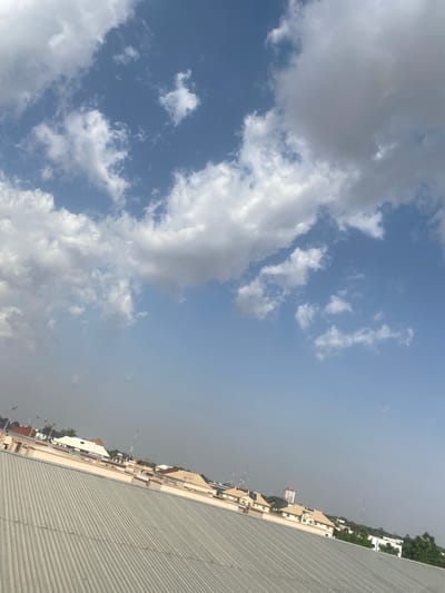 The image depicts an outdoor scene in Fagge A, Nigeria, likely during the daytime given the bright sky. The composition is dominated by a dynamic sky filled with fluffy, white cumulus clouds against a backdrop of a clear blue sky. The clouds are scattered and appear to be moving, suggesting a breezy day.
On the right side of the frame, the perspective shifts to a view of buildings and structures. A corrugated metal roof takes up a significant portion of the foreground, angled upwards. Beyond the roof, several buildings of varying heights and architectural styles are visible, with some featuring light-colored facades and windows. Trees with lush green foliage dot the landscape behind the buildings, indicating a warm climate. In the distance, what appear to be telecommunication towers or antennas rise above the structures. The hazy atmosphere suggests it might be a warm or humid day, or possibly dust in the air. There is no discernible activity or event taking place; the scene appears to be a static view of the environment. No people are visible in the image, and there is no visible text. The overall impression is one of a typical urban or semi-urban environment in a tropical region under a cloudy sky.