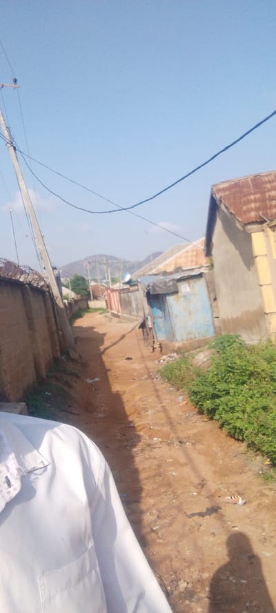 The image depicts a dirt road in a residential area in Kwamba, Nigeria. The road is unpaved and appears to be in a state of disrepair, with visible debris and rough patches. On either side of the road, there are buildings constructed with corrugated metal roofs and various wall materials, including concrete blocks and what appear to be makeshift structures. A tall utility pole with wires runs along the left side of the road, and a barbed wire fence can be seen along the wall. In the background, a hill can be seen, suggesting a somewhat hilly terrain. A person, partially visible in the foreground, is wearing a white collared shirt. The overall impression is of a developing or informal urban settlement.