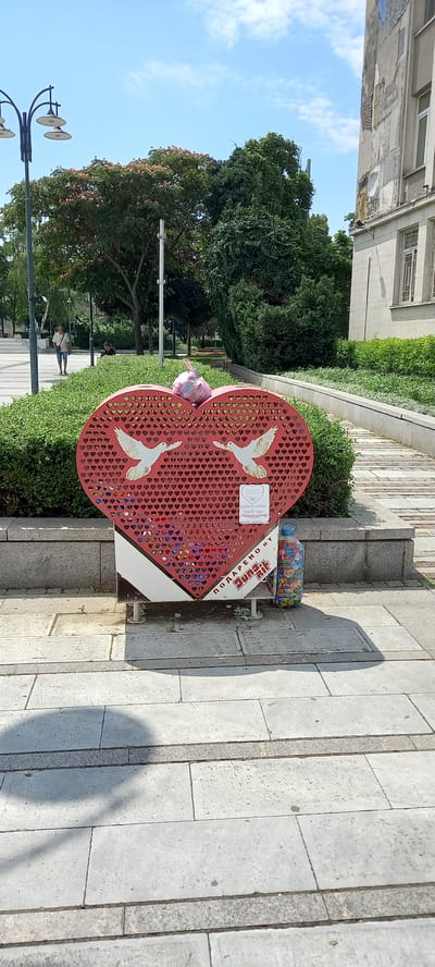 In Silistra, Bulgaria, a large, stylized red heart sculpture sits in a public space. The heart is covered in a pattern of small, cut-out hearts and features two white doves in flight on its surface. Below the doves, a small sign is attached. The base of the sculpture is white and extends to the ground. A large plastic bottle filled with colorful bottle caps is placed to the right of the sculpture. The sculpture is situated in a paved area with a backdrop of manicured green bushes and trees. In the background, a lamppost, a building with peeling paint, and a few individuals walking are visible. The scene suggests an outdoor urban setting, possibly a park or plaza.