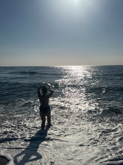 The image shows a person from behind, standing at the edge of the ocean, with waves washing over their feet. The person is wearing a dark bikini and has their hands up, touching their hair. The sun is bright and reflecting off the water, creating a shimmering effect. The sky is a clear blue, with a slight haze near the horizon. The overall scene is one of a serene beach setting. The location is not specified as Abang, Indonesia.