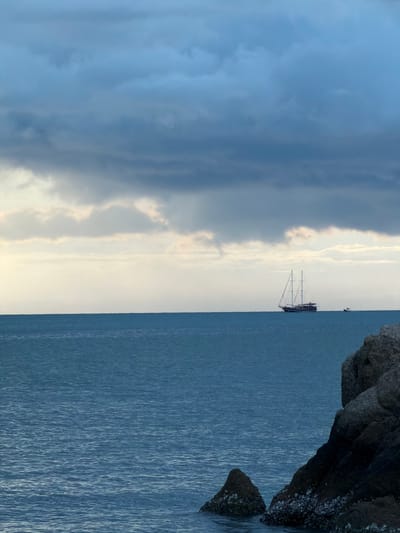 A wide shot captures a serene seascape with a sailboat sailing on the horizon, set against a dramatic cloudy sky. The water is a deep blue, with gentle waves creating a rippled texture. In the foreground, large, dark rocks are visible on the right side of the frame, with smaller rocks and shells emerging from the water. The sailboat, with its sails extended, is positioned in the middle of the frame, appearing small against the vastness of the ocean and sky. The sky is filled with heavy, dark clouds that are beginning to break, allowing patches of lighter sky and hints of sunlight to peek through, creating a moody and atmospheric scene. A smaller boat is visible to the right of the sailboat. The overall impression is one of peaceful solitude and the power of nature. This scene could be located in Baan Bo Phut, Thailand, known for its beautiful beaches and coastal views.
