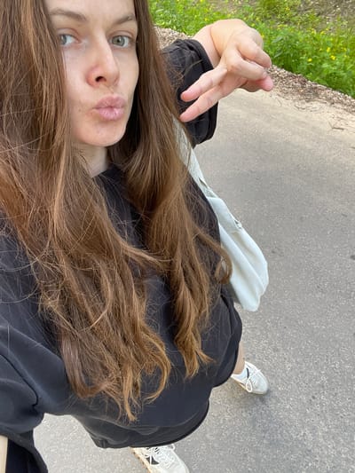 The image shows a selfie taken from a high angle, looking down at the subject and the surrounding environment. The subject is a young woman with long, brown hair that cascades over her shoulders and partially obscures her face. She has light green eyes and is wearing a black outfit. Her lips are pursed in a playful expression. She is holding up her left hand with two fingers extended, forming a peace sign.
In the background, a paved path is visible, suggesting an outdoor setting. To the left of the path, there is a patch of green grass with some yellow flowers, indicating a natural environment, possibly a park or a roadside. A white bag is placed on the ground next to the subject. Her white sneakers are also visible, suggesting she is standing or walking on the path.
The lighting suggests it is daytime, with soft natural light. The overall mood of the image is casual and cheerful. The location is identified as Zarechny, Russia. There is no visible text in the image.