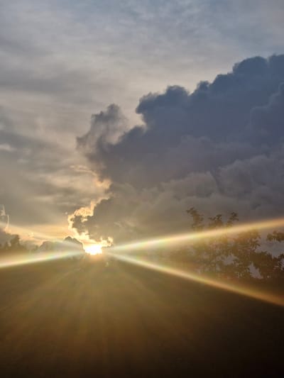 This photograph captures a dramatic sunset over the city of Saulgoži, Latvia. The sky is filled with a mix of dark, heavy clouds and thinner, lighter ones that are illuminated by the setting sun. Beams of golden light are radiating from behind a cluster of clouds, creating a powerful visual effect. The lower portion of the image is dominated by a silhouetted landscape, possibly featuring trees or buildings, and a hazy foreground that suggests a field or body of water. The overall mood is one of intense natural beauty and a sense of transition as day turns into night.