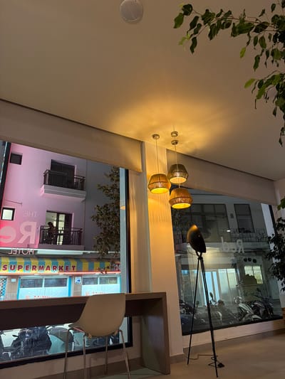 The image shows an interior view from a cafe or restaurant looking out onto a street in Sant Antoni De Portmany, Spain. The cafe features a long counter with a white chair and a floor lamp with a black shade. Outside, a building with pink and white signage that reads "THE RC HOTEL" is visible, along with a "SUPERMARKET" sign. Several motorcycles are parked in front of the supermarket. The lighting suggests it is evening or nighttime.