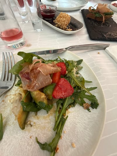 An overhead, close-up shot shows a partially eaten salad on a white, textured plate. The salad consists of arugula, sliced strawberries, avocado, prosciutto, and what appear to be bell peppers and croutons. A fork and knife rest on the plate. In the background, a glass of pink liquid, two empty shot glasses, and a small dish of what appears to be jam or preserves are visible. A slate platter with a small appetizer is also in the background. The scene is set on a white tablecloth with a subtle, woven pattern. The lighting is bright and appears to be natural, casting soft shadows. The overall impression is that of a casual dining setting. There is no visible indication of the city of Pavlohrad, Ukraine, or any specific details that would place the image there.