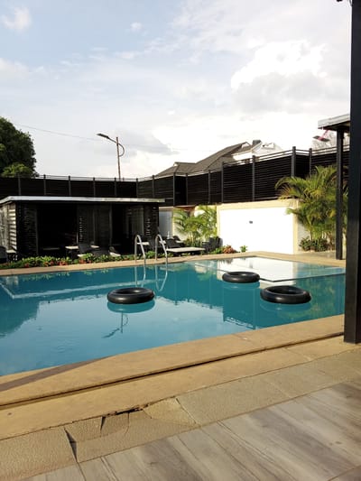 A rectangular swimming pool is filled with clear blue water and has three black inflatable rings floating in it. The pool is surrounded by a light-colored tiled deck and a raised concrete edge. In the background, there is a black structure with horizontal slats, possibly a covered seating area, with chairs and tables visible inside. Beyond that, there is a black slatted fence and palm trees. Above the fence, rooftops of buildings and a streetlamp are visible. The sky is partly cloudy with white clouds against a pale blue sky. The lighting suggests it is either early morning or late afternoon. The scene appears to be a tranquil setting, likely a resort or private residence in Unguwar Sarki, Nigeria.