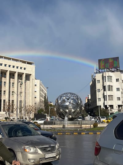 This image depicts Yousef Al-Azma Square in Sarouja Municipality, Damascus, Syria, on a clear day following rain, evidenced by wet pavement and a visible rainbow. The scene is dominated by a large, multifaceted, reflective metallic spherical sculpture (the Sarouja Globe) positioned within a circular fountain roundabout in the center. Water jets are actively spraying from the fountain.

To the left of the sphere stands a multi-story, light-colored building with classical architectural features, identified as the Syrian Ministry of Justice, displaying multiple Syrian flags on flagpoles. To the right of the sphere, a multi-story building with numerous windows features a large, colorful electronic billboard on its rooftop, along with various antennae.

A prominent rainbow arcs across the upper portion of the light blue sky, which contains scattered white clouds. A small dark bird is visible in flight beneath the rainbow on the left side.

The foreground shows wet asphalt. A light gold Kia Cerato sedan with a Syrian license plate (389330) is partially visible in the lower left, with a person's arm partially obscuring its lower corner. The rear of a silver SUV is visible on the far right. Multiple other vehicles, including white cars, dark sedans, and yellow taxis, are present on the wet roadway around the roundabout. Several pedestrians are visible on the sidewalk along the right side of the roundabout. Trees and streetlights are interspersed throughout the urban environment.