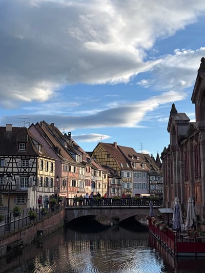 A vertical, eye-level medium shot captures a picturesque canal scene in Colmar, France, under a dramatic sky filled with textured clouds and patches of blue. The canal, reflecting the light, flows from the foreground toward the background, flanked by quaint, colorful half-timbered buildings. On the left bank, a stone wall lined with lampposts and hanging flower baskets separates the canal from a walkway. Several people are visible on this walkway, some walking, others standing and conversing. A brick bridge with an arched underside spans the canal, its top serving as a pedestrian walkway. More people are gathered on the bridge, observing the scenery. On the right bank, a building with large arched windows and intricate stonework dominates the right side of the frame. In front of this building, a red-railed outdoor seating area with tables, chairs, and closed umbrellas suggests a cafe or restaurant. The architecture of the buildings is characteristic of Alsace, with steeply pitched roofs and decorative half-timbering. The overall impression is one of a charming, historic European town.