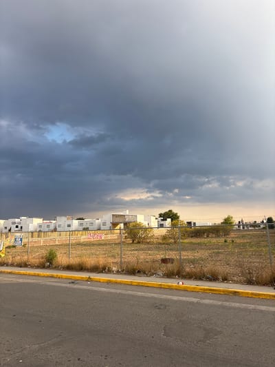 The image depicts a view along a road in San Juan Cuautlancingo, Mexico. A line of white buildings, likely residential, stands behind a metal fence. Between the fence and the road is a strip of dry, grassy land. The road itself is asphalt, marked with a yellow line. The sky is overcast, with dark storm clouds dominating the view.