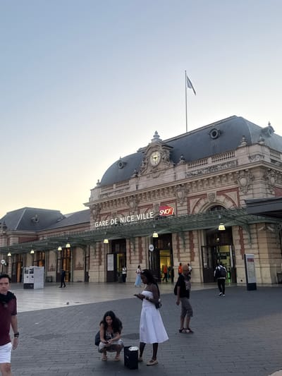 The image displays the exterior of Nice-Ville railway station in Nice, France, at what appears to be dusk or dawn. The station's ornate, classical architecture features a light-colored stone facade with reddish-brown brick accents, decorative cornices, pilasters, and arched entryways. A prominent clock tower on the central section of the roof shows the time as approximately 7:45. The roof is a dark, possibly slate, mansard style with several dormer windows. A plain light-colored flag flies from a flagpole on the roof above the main entrance.
A large, glass and metal canopy extends over the main entrance, supported by intricate metalwork, with illuminated pendant lights hanging beneath it. The signage "GARE DE NICE VILLE" is prominently displayed in white, illuminated letters above the canopy, accompanied by the red and white "SNCF" logo.
The foreground is a paved plaza of alternating light and dark grey rectangular tiles. Numerous individuals are present on the plaza. On the far left, a man wearing a maroon shirt, black scarf, and white shorts is walking towards the left of the frame. In the center-left, a woman with dark hair, dressed in a patterned dress, is squatting and looking at a mobile phone, with a dark bag beside her on the ground. To her right, a woman in a white strapless dress stands while holding a mobile phone; she has a dark bag on her shoulder. Further right, a man in a black t-shirt, light shorts, and sneakers is walking towards the left.
In the midground, near the station entrances, several other individuals are visible. One person with a white backpack is entering a doorway labeled "Hall 1" on the right. Other people are seen walking or standing near the various entrances. The sky above is a gradient of light blue transitioning to a pale orange-yellow near the horizon, indicating the time of day. Artificial lights under the canopy provide illumination to the immediate area in front of the station.