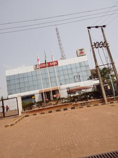 The image displays the exterior of a Zenith Bank branch located in Ankpa, Nigeria, under a clear sky. The two-story building features a large "ZENITH BANK" sign in red lettering with a "Z" logo on its upper facade, which is predominantly composed of reflective glass panels. A smaller "Z ZENITH" logo cube is situated on the roofline to the right. Three flagpoles stand in front of the building; one bears a green and white flag.
The bank premises are secured by a black metal fence. An open gate on the left side of the fence provides access. A paved area of reddish-brown interlocking bricks forms the foreground, bordered by a curb with black and white stripes.
Two individuals are present: one person in a red shirt is partially visible through the open gate on the left, near the bank's entrance. Another person wearing a pink shirt is faintly visible behind the fence and shrubs, closer to the building. A white vehicle, possibly an SUV or minivan, is parked behind the fence, visible through the lower glass facade.
In the background, a tall lattice antenna tower is visible to the left of the building. Utility poles with overhead wires are prominent on the right side of the frame. Green foliage is visible behind the fence and near the utility poles.