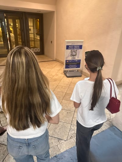 In Coral Gables, Florida, two individuals are walking away from the camera in an indoor hallway with tiled flooring and beige walls. A sign with a QR code and the text "MUST PREPAY UPON ARRIVAL" is visible on the right side of the image. The person on the left has long, blonde-highlighted hair and is wearing a white t-shirt and light blue jeans. The person on the right has dark brown hair tied in a ponytail, sunglasses on their head, and is wearing a white t-shirt, dark pants, and carrying a red handbag. In the background, there are double glass doors leading to an area with visible interior lighting and reflections.