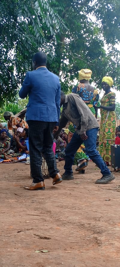 The image depicts an outdoor gathering in Fizi, Democratic Republic of the Congo, taking place on an unpaved, reddish-brown dirt ground under the canopy of green-leafed trees. In the foreground, a male subject is visible from the back, wearing a blue blazer, dark plaid trousers, and brown dress shoes, standing upright and facing away from the camera. To his right and slightly forward, another male subject is bent significantly at the waist, facing towards the right side of the frame. This individual wears a brown striped long-sleeve shirt, blue jeans, and dark, thick-soled athletic shoes.
In the midground and background, an assembly of people, predominantly women, is observed. Many women are seated on the ground, dressed in various brightly patterned African print fabrics and head coverings, some appearing to clap or gesture with their hands. Behind the bent-over male, two women stand; one wears a vibrant multi-colored patterned dress with a yellow headwrap, and the other a green and yellow floral patterned dress with a matching yellow headwrap. A black speaker or sound equipment is partially visible on the far right edge of the frame. The overall scene suggests a community or public event.