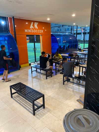 A group of young men are gathered at an indoor mini-soccer facility. The setting appears to be a recreational or sports center, with brightly lit indoor fields visible in the background. The overall ambiance suggests a casual, possibly after-school or weekend gathering.
The primary subjects are several young men, mostly dressed in athletic wear. Some are seated on black metal benches, observing or resting, while others are standing or walking around the facility. One person in the foreground is wearing a black t-shirt and shorts with red stripes, and another is in a black t-shirt and shorts. A person in a yellow shirt is also visible.
The activity taking place is likely related to indoor soccer or a break from playing. The presence of benches and the general layout suggest a space designed for players to relax or wait between games. The lighting is bright and even, indicating an indoor environment.
Notable details include the orange wall with white text that reads "AK MINISOCER". The blue translucent structures add a modern and vibrant touch to the space. The floor is tiled with light-colored square tiles. The time of day cannot be determined definitively due to the indoor setting, but the atmosphere is energetic and youthful. There are no clear indications of the weather or specific emotions, though the scene appears relaxed. The presence of a large grey trash bin in the upper right corner suggests a functional aspect of the facility.