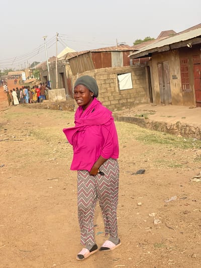 A woman in a pink shirt and patterned pants stands on a dirt ground with buildings in the background. She is wearing a gray head covering and light-colored sandals with socks. A crowd of people stands in the distance. The buildings are constructed from various materials, including brick and corrugated metal. The overall atmosphere is casual and rural. The woman appears to be looking toward the viewer.