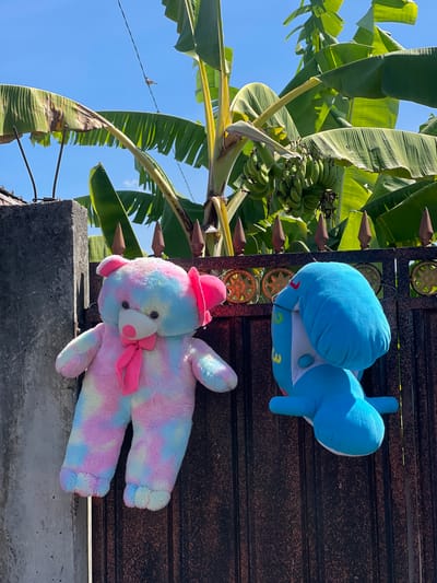 A large, tie-dyed teddy bear in pink, blue, and yellow hues, adorned with a pink bow, hangs from a metal fence. To its right, a blue stuffed dolphin is also suspended from the same fence. The fence is ornate with decorative circular elements and pointed finials along the top. Behind the fence, a banana tree with a cluster of green bananas is visible against a bright blue sky. The scene appears to be an outdoor setting, possibly in a residential area of Sukawati, Indonesia, indicated by the presence of a concrete pillar and the tropical vegetation. The lighting suggests it is daytime.