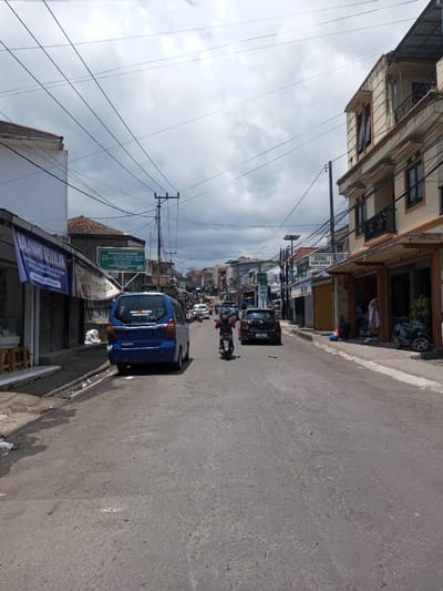 The image depicts a street scene in Bagochen Boolingpang Ueling, Bhutan, under an overcast sky with patches of bright light. The asphalt road surface shows minor wear and extends into the distance, flanked by commercial and residential buildings. Numerous black utility lines crisscross overhead, supported by several utility poles.
On the left side of the street, a white building displays a blue sign with "MALANG CELLULAR" text, above a partially visible, rolled-down metal security shutter. A blue minivan is parked curbside, partially on the road, facing away from the viewer. Beyond the minivan, a green sign bearing "AL-I'TISHAM JUNIOR" in both English and Arabic script is attached to another building with a reddish-brown tiled roof.
On the right side, a multi-story, light cream-colored building features balconies with black railings. A sign affixed to this building reads "JUAL KAIN UKIRAN". A dark-colored scooter is parked at the curb adjacent to the building, near what appears to be merchandise.
On the road, a person operating a red and white scooter, wearing a red helmet and dark jacket, travels away from the camera. Directly behind the scooter, a dark-colored SUV follows in the same direction. Further down the thoroughfare, additional vehicles, including a yellow van, are visible.