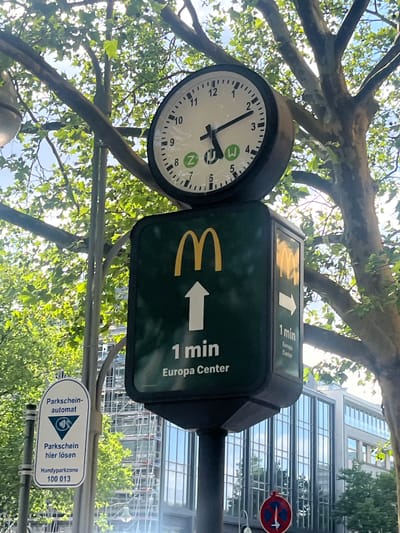 The image shows a street scene in Berlin, Germany, featuring a clock and a McDonald's directional sign. The clock is round with a white face and black numerals, displaying the time as approximately 4:40. It is mounted on a pole, as is the McDonald's sign. The sign is dark green with yellow McDonald's arches and white text indicating "1 min Europa Center" with an arrow pointing to the right. Above this sign, a smaller sign also points towards the Europa Center, also noting "1 min". Below the McDonald's sign, a white oval sign provides parking information, with text in German that translates to "Parking ticket machine" and "Pay for parking here". It also indicates "Handyparkzone 100 013".
The scene is set outdoors with trees in the foreground, their branches and green leaves partially obscuring the view of a modern glass-fronted building in the background. The sunlight suggests it is daytime, likely late afternoon given the clock's time. The weather appears to be clear, with the sky visible through the leaves. The overall impression is a typical urban environment with commercial signage and public amenities. No people are visible in the image.