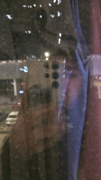 The image shows a blurry reflection inside a car window, likely at night. Visible through the reflections are city lights and buildings. A device with several circular openings, possibly a camera or electronic gadget, is also reflected in the window, along with what might be the driver's head silhouette. The window itself has horizontal defroster lines visible.