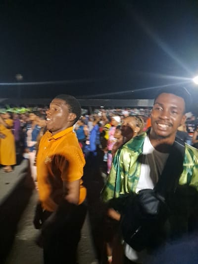 The image captures two young men posing for a photo at an event, likely in Owerri, Nigeria. The man at the top wears a green and white patterned top over a white t-shirt with a black bag slung across his shoulder. The man at the bottom is wearing an orange collared shirt. In the background, a large crowd is gathered, suggesting a public event. A streetlamp illuminates the scene. The ground is asphalt.