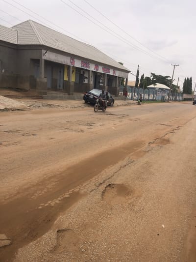The image shows a street scene in Hanwa, Nigeria. The road is unpaved, with significant damage and potholes. A building with a light-colored roof and painted murals is visible along the side of the road. A dark-colored car is parked near the building, and a motorcycle with two riders is positioned next to it. Several people are walking along the sidewalk in the distance. Overhead electrical wires are visible. The sky is overcast.