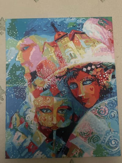 This is a photograph of a painting. The painting is an abstract depiction of what appear to be three faces, possibly women, rendered in a vibrant and expressive style. The faces are intertwined and overlap, creating a sense of depth and movement. The color palette is rich and diverse, featuring blues, oranges, reds, yellows, pinks, and white. There are various patterns and textures within the painting, including dots, swirls, and checkered elements, adding to its abstract and artistic nature. The faces have distinct features like eyes and noses, but they are stylized and not anatomically precise. The background is also abstract, with a mix of swirling patterns and solid colors. The overall impression is one of energy and emotion, possibly representing a dreamlike or surreal scene. The painting is displayed on a wall with a textured wallpaper. There is no visible text in the image.