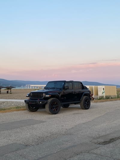 A black Jeep Wrangler Rubicon is parked on an asphalt road. The vehicle is positioned facing slightly left of the camera, with its front end closer. The Jeep has custom black rims with red accents. In the background, there is a sandy beach with several umbrella-like structures and a white, open-air pavilion. Beyond the beach, there are rolling hills and mountains, and the sky is a gradient of soft blue, pink, and pale yellow, suggesting either sunrise or sunset. To the right of the Jeep, there is a beige building with posters on it, one featuring an image of a woman. The scene appears to be a coastal area, possibly near the city of Novorossiysk, Russia, given the context.