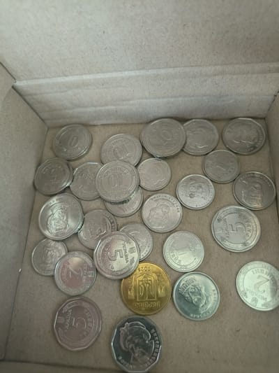 The image shows a collection of Ukrainian coins scattered inside a cardboard box. The coins are of various denominations, including 10, 5, 2, and 1 hryvnia. Some coins feature portraits of historical figures, while others have national symbols. The lighting suggests an indoor environment. The location in Kalynivka, Ukraine, is not discernible from the image itself. The scene is a still life, with no people, actions, or interactions depicted.