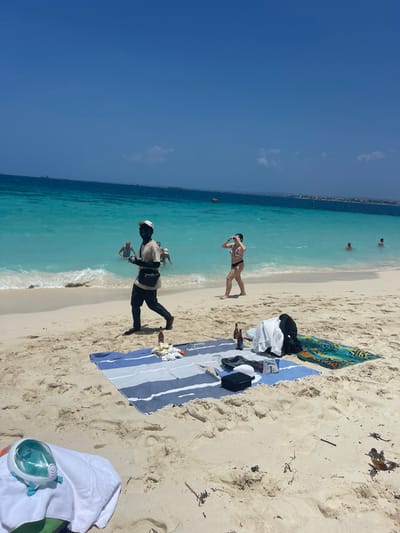 The image depicts a sunlit beach scene in Zanzibar, Tanzania. The sky is clear and blue with sparse white clouds. The ocean water transitions from light turquoise near the shore to deeper blue further out, with small waves breaking on the sandy coast.
In the mid-ground, a dark-skinned male, wearing a light-colored long-sleeved shirt, dark trousers, dark shoes, and a dark baseball cap, walks from left to right across the sand. He carries several small, light-colored figures in his left hand, holding them up, and a dark cross-body bag hangs from his shoulder. He looks directly forward, towards the viewer. To his right, a light-skinned female in a dark two-piece bikini walks out of the shallow water onto the sand, her arms slightly raised. Further in the water to the right, three other individuals are visible swimming or wading. A yellow buoy floats in the mid-distance water.
On the foreground-midground sand, a blue and white striped beach towel is spread. It holds two brown glass bottles, a crumpled white item, a white garment, a pair of dark shoes, and a small black rectangular container. Adjacent to this towel, a colorful patterned beach mat is laid out. In the lower-left foreground, a white and light blue full-face snorkeling mask rests on a white towel. The light-colored sand exhibits footprints and scattered dark organic debris. On the distant horizon, a landmass with several visible structures and multiple vessels are present.