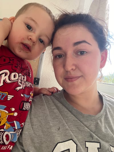 This is a selfie of a woman and a baby. The woman is in the foreground, her face tilted towards the camera and slightly out of focus. She has dark hair and is wearing a grey t-shirt. To her left, and slightly lower, is a baby. The baby has light brown hair and big, dark eyes. The baby's face has some redness and what appears to be food residue around its mouth. The baby is wearing a red shirt with a graphic print of what looks like cartoon characters and text. The visible text on the baby's shirt reads "Roaring Sport".
The setting appears to be indoors, with a window in the background suggesting daylight. The overall lighting is bright, indicating it's daytime. There are no explicit cues to suggest the weather or a specific location within Chelyabinsk, Russia, beyond the provided context. The image conveys a sense of intimacy and a casual moment between the woman and the baby. The woman's expression is soft, and the baby looks directly at the camera.