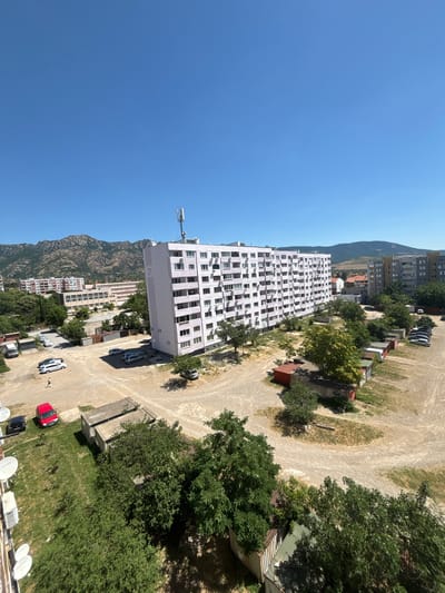 The image depicts an aerial view of a residential area in Sliven, Bulgaria, on a clear, sunny day. The foreground is dominated by lush green trees and grass, with satellite dishes visible on the side of a building. Moving further back, a large, pale pink multi-story apartment building with numerous balconies stands prominently. Adjacent to this, several smaller, less distinct residential buildings and a long, beige building that appears to be a school or administrative center are visible. In the background, rugged, rocky mountains rise against the bright blue sky. The ground in front of the apartment buildings is a dirt lot with several parked cars and a few small, red-roofed structures that could be garages or storage units. A single individual can be seen walking across the dirt lot. The overall scene suggests a typical urban residential environment in a region with mountainous topography.
