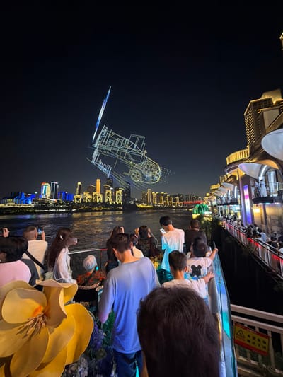 The image depicts a nighttime scene along a waterfront, likely in Nan'an District, China, given the distinctive illuminated cityscape in the background. A group of people are gathered on what appears to be a boat or elevated viewing platform, facing a river that separates them from the city. The city skyline is a prominent feature, with numerous buildings illuminated in various colors, including blue, yellow, and white. A particularly striking element in the sky above the city is a complex drone light show, forming geometric shapes and patterns that are brightly lit against the dark sky.
In the foreground and midground, the focus is on the people observing the spectacle. They are diverse in age and gender, with many looking towards the city and the drone show. Several individuals are holding up smartphones, presumably to capture photos or videos of the event. The people are casually dressed, suggesting a leisure outing. A large, decorative yellow flower is partially visible in the bottom left corner of the frame, adding a decorative element to the viewing area. The water of the river is visible, reflecting the city lights. The right side of the image shows part of a modern building structure with balconies and walkways, also illuminated and occupied by people, suggesting a commercial or entertainment area. The overall atmosphere is one of awe and engagement with the visual display.