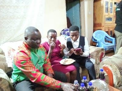 Three individuals are sitting on a couch. The person on the left wears a colorful green and red patterned shirt. In the middle is a person in a red dress eating. The person on the right is dressed in a white shirt with a black vest and pants, looking at their phone. A plate of food sits on their lap. Several bottles with blue caps are on the table in front of them. In the background, there's a blue plastic chair and a wooden cabinet.