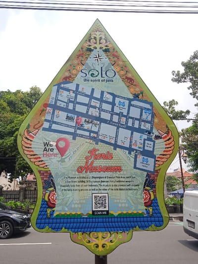 The image shows a signpost in Surakarta, Indonesia, located at coordinates -7.569, 110.811. The signpost is a triangular shape with a bright blue background. The signpost features the words "Solo the spirit of Java" with a stylized compass rose in the center. Beneath this, there is a map of the city of Surakarta, with a red pin pointing to the location of "Keris Museum." The sign also indicates the museum's location at "Jl. Bhayangkara at Sriwedari Park Area," and that it has "four floors building." Beneath the map, there is a description of the museum, noting that it has "many traditional weapons, especially keris from all over Indonesia." It also boasts a "video of the keris history in Indonesia" and the "keris making process." A QR code is present, with the words "SCAN ME" beneath it. The sign is in front of a partially obscured view of a street, with trees and buildings. The image captures a vibrant blend of local culture and historical interest.