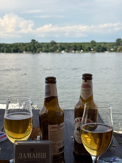 The image depicts a serene outdoor setting by a body of water, likely the Danube River, given the context of Zemun Urban Municipality, Serbia. In the foreground, on a wooden table, are two glass bottles of Birra Moretti beer and two glasses partially filled with the same beverage. The labels on the bottles are clearly visible, indicating "BIRRA MORETTI" and "THE ITALIAN PASSION FOR BREWING." One of the glasses has a foamy head. To the left, partially visible on the table, is a small box with Cyrillic text, possibly containing cigarettes or matches. The background features a wide expanse of water under a cloudy sky. Across the water, a dense green treeline borders the horizon, with some small structures and boats subtly visible amongst the trees. The overall impression is one of a relaxed moment, possibly a break at a riverside cafe or restaurant. No people are visible in the image.