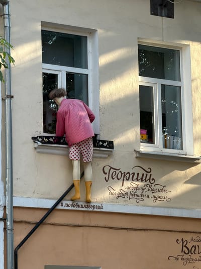 The image shows a quirky art installation on the exterior wall of a building in Brest, Belarus. The main subject is a mannequin dressed in a pink striped jacket and heart-patterned shorts, with yellow boots. The mannequin appears to be positioned as if it's leaning over a window ledge, with its head and upper body inside or reaching into the window.
To the right of the mannequin, on the beige wall, is an inscription in stylized brown lettering. The text reads "Теории любовник Ваулавы" which translates to "Theories of a lover Vavlava." Below this, there is another line of text, harder to decipher, that appears to be "своя убил ревнивым мужем" which might translate to "his own killed by a jealous husband." Further to the right, there is more text in a different font, starting with "Малодо" and then "Вау," and "не выдержала," which can be translated as "young" "Vav" and "couldn't stand it." Below this, there is a hashtag: "#любовникгеоргий" meaning "#lovergeorgiy."
The scene appears to be outdoors, with natural sunlight casting shadows on the wall, suggesting it is daytime. The building has two windows visible, with one appearing to contain stacks of papers or books. The overall setting is urban, with visible pipes running along the wall and a drainpipe at the bottom of the image. The art installation, with its unusual subject and inscribed text, creates a surreal and perhaps narrative-driven visual.