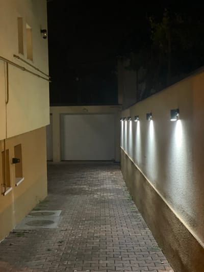 The image presents an outdoor view, potentially a public or semi-public space in Sremska Kamenica, Serbia, at night. The viewpoint is low, looking upward.
Overhead, a concrete overhang is illuminated by a row of evenly spaced, directional lights. The pavement alongside the overhang is constructed with interlocking concrete pavers. The wall is brick.
Below the overhang, there's a mix of architectural elements. On the left is a building or structure with a light-colored facade and what appear to be pipes running along the exterior. There are two square openings. On the right, the structure continues with similar materials. Two round vent openings are visible. A closed garage door is in the background.
The scene is dark, with the artificial lights being the primary source of illumination. There are no people visible.