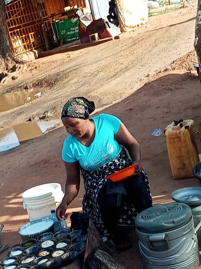 In Wuro Dole, Nigeria, a woman in a bright blue t-shirt and a patterned skirt is preparing food outdoors. She wears a headscarf adorned with reflective embellishments. In front of her is a tray filled with small, round, cooked items, possibly cakes or dumplings, arranged in a grid. She is holding an orange tray, and a large white bucket sits to her left. The ground is dirt, with some puddles of water, suggesting a natural environment. In the background, a stall with orange bars is visible, with a green bag bearing lettering in what appears to be an African script. There are also some containers and possibly other food items on display. The scene depicts a street food vendor at work in a bustling outdoor setting.