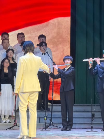 An indoor musical performance or rehearsal is captured on a brightly lit stage in Guta, China. The image features a group of musicians, primarily dressed in formal attire. In the foreground, a person wearing a bright yellow suit stands with their back to the viewer, holding and playing a flute. Immediately behind them, another individual in a black suit with short, dark hair and glasses is playing a flute, facing towards the camera. Further back and to the left, another person in a black suit is also seen playing a flute.
Several other performers are visible behind and to the sides of the main flutists. Most of these individuals wear white shirts, and some have glasses, appearing focused on the activity. At the bottom right, a few performers in black tops and white skirts or dresses are also part of the ensemble.
The stage is equipped with multiple microphones on stands, positioned in front of the musicians. The backdrop on the right side of the stage is a vibrant red, possibly a large screen or fabric curtain, while the left side features a darker, possibly corrugated, wall structure. The floor appears to be a light-colored stage. The overall scene suggests a concert or formal musical event, with performers concentrating on their instruments. No visible text is present in the image.