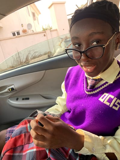 A young person with black glasses and a headband is indoors, seemingly seated in a car. They wear a white collared shirt, a purple sweater vest with white lettering, and plaid shorts. The car door panel is visible, as are some buildings outside the window. They are angled towards the camera, smiling slightly. One hand rests in their lap, near the car door.